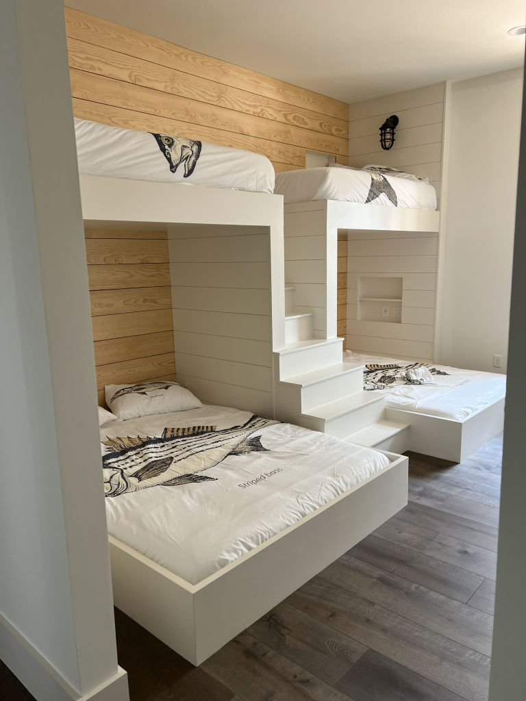 Built-in white bunk room with four beds, integrated staircase to upper bunks, shiplap walls, wall sconces, and dark wood flooring.